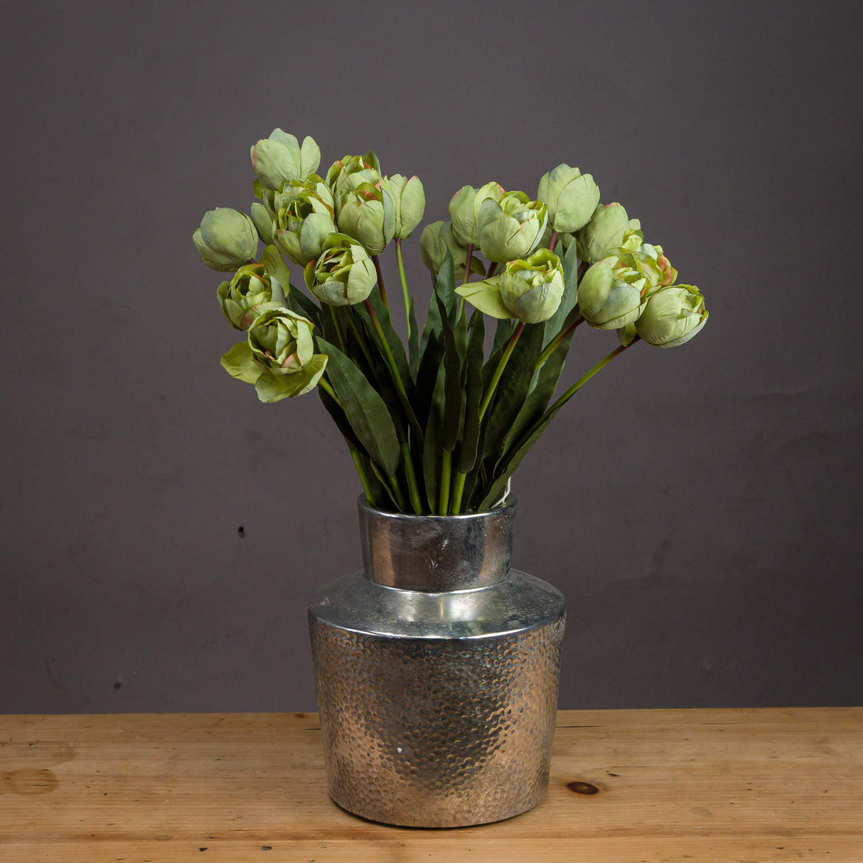 Hill Interiors Green Tulip faux stems arranged in a hammered silver metal vase on a wooden surface against a dark grey background.