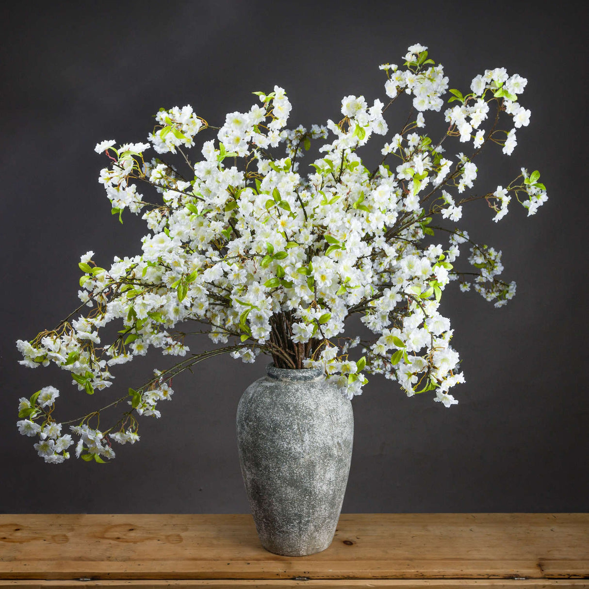Hill Interiors White Cherry Blossom Spray arranged as a full display in an aged stone vase on a wooden surface against a dark grey background.