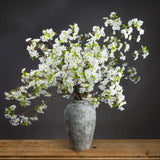 Hill Interiors White Cherry Blossom Spray arranged as a full display in an aged stone vase on a wooden surface against a dark grey background.