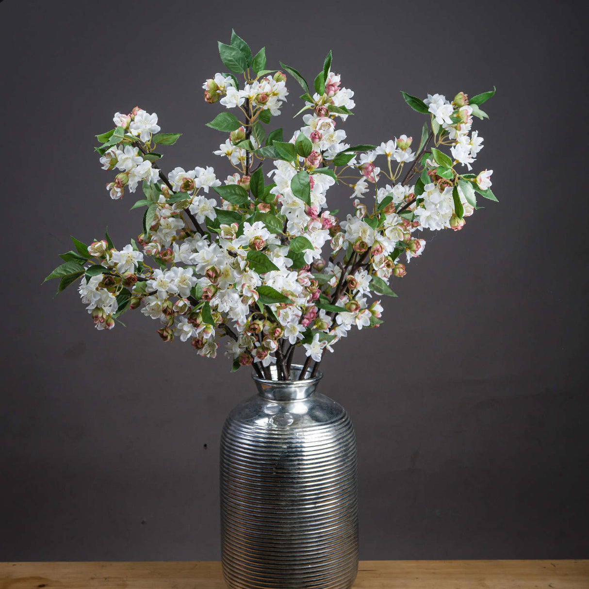 Hill Interiors English Blossom Branch faux stems with white flowers, pink buds and green leaves arranged in a ribbed silver vase against a dark grey background.