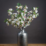Hill Interiors English Blossom Branch faux stems with white flowers, pink buds and green leaves arranged in a ribbed silver vase against a dark grey background.