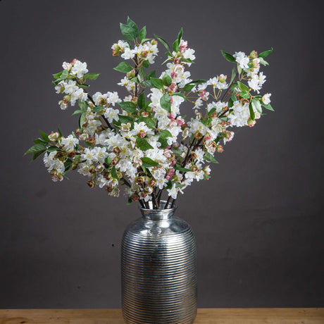 Hill Interiors English Blossom Branch faux stems with white flowers, pink buds and green leaves arranged in a ribbed silver vase against a dark grey background.