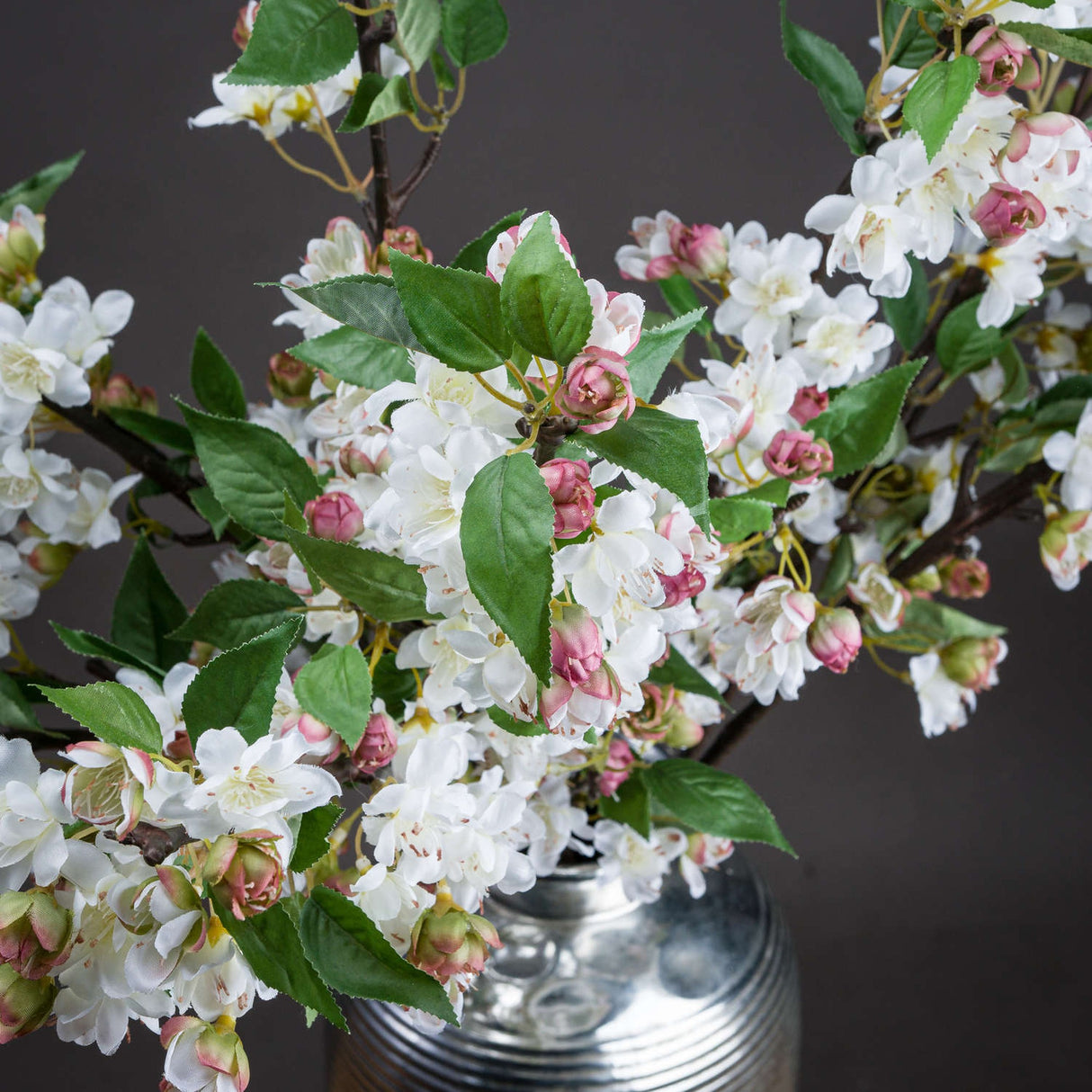 Hill Interiors English Blossom Branch faux stem with white flowers, pink buds and green leaves on brown branches, arranged in a silver vase.