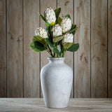 Hill Interiors Closed White Protea faux stems arranged in a distressed white ceramic vase on a wooden surface against a rustic timber plank backdrop.