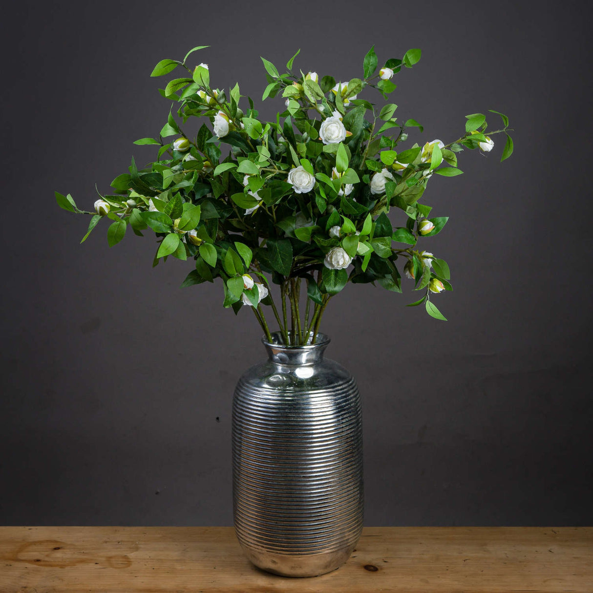 Hill Interiors Cream Gardenia Stems arranged in a ribbed silver vase on a wooden surface against a dark grey background.