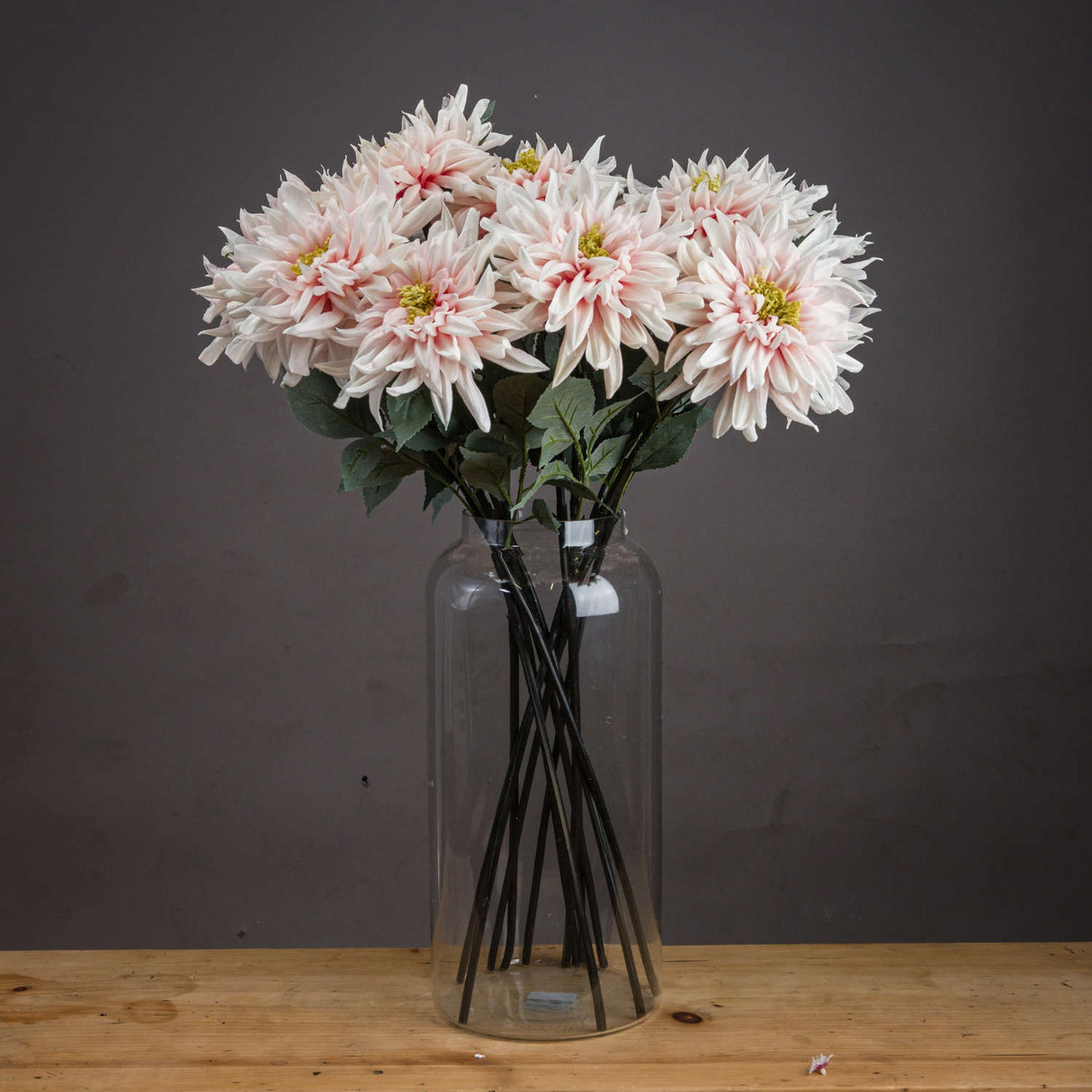 Hill Interiors Lush Pink Dahlia faux stems arranged in a tall clear glass vase on a wooden surface against a dark grey background.