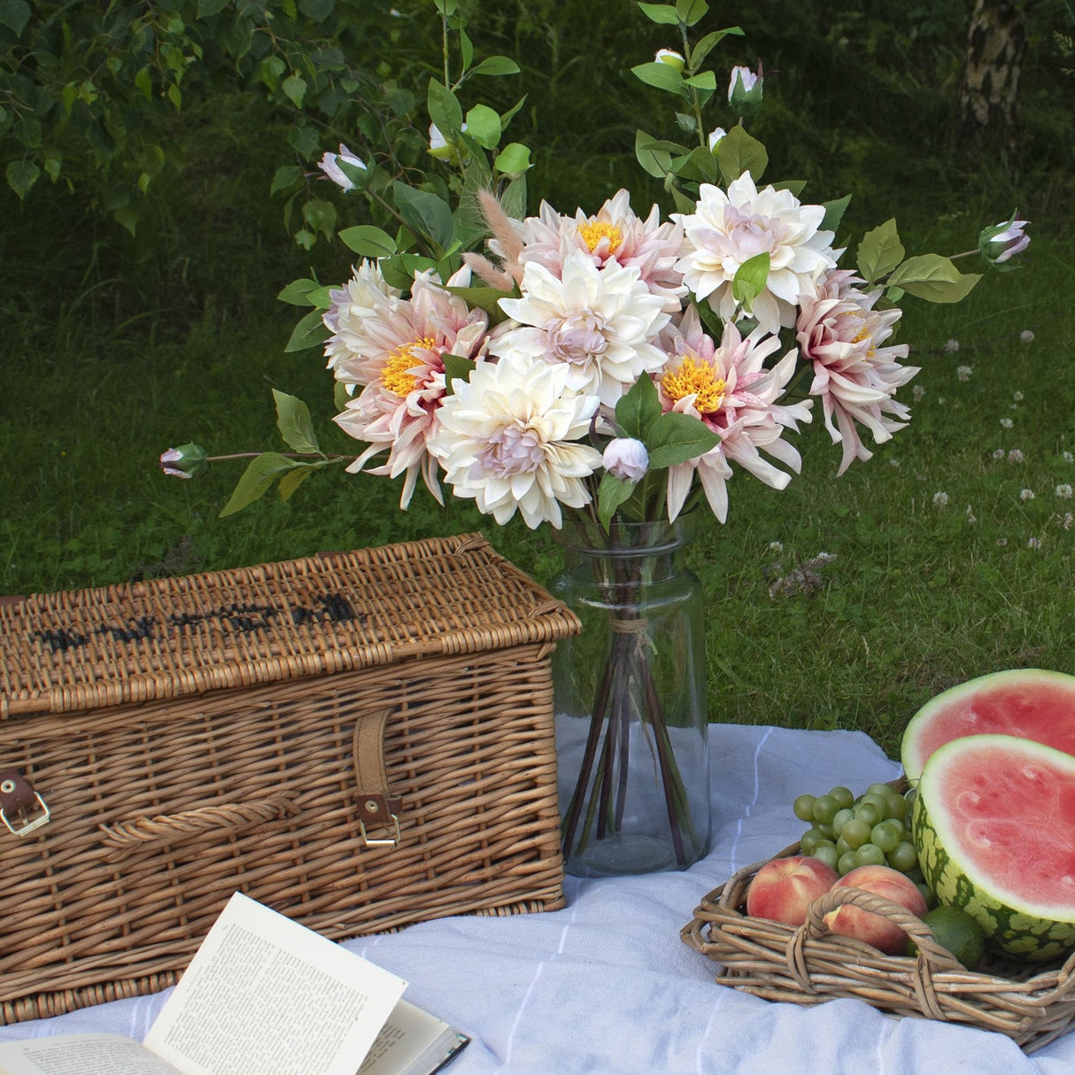 Hill Interiors Lush Pink Dahlia faux stems arranged with foliage in a glass vase beside a wicker picnic hamper, book and fruit tray on garden grass.