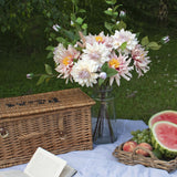Hill Interiors Lush Pink Dahlia faux stems arranged with foliage in a glass vase beside a wicker picnic hamper, book and fruit tray on garden grass.