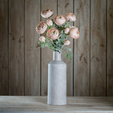 Hill Interiors Pastel Pink Ranunculus faux stems with ruffled blooms and green foliage arranged in a tall stone-grey bottle vase on a wooden surface.