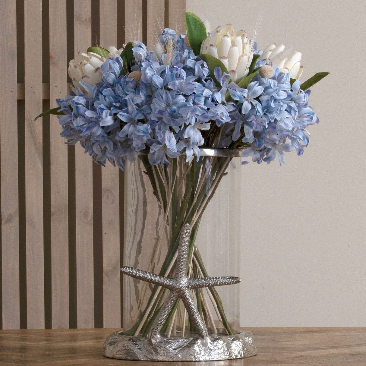 Hill Interiors Light Blue Large Headed Agapanthus faux stems arranged with white proteas in a glass vase with silver starfish base on a wooden table.