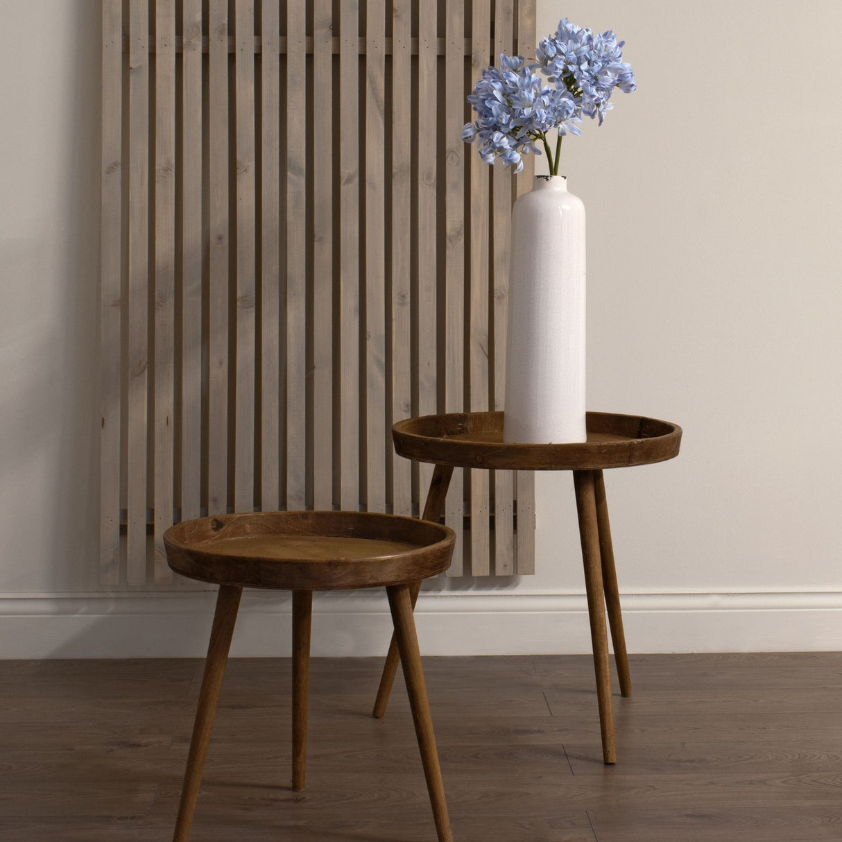 Hill Interiors Light Blue Large Headed Agapanthus stem in a tall white ceramic vase on a round wooden side table beside a slatted wood wall panel.