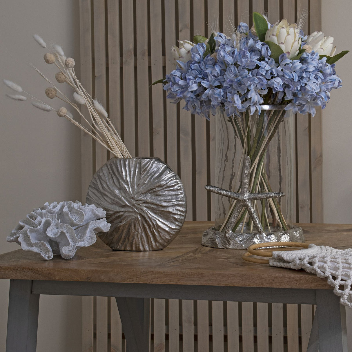 Hill Interiors light blue large headed agapanthus arranged with white proteas in a glass vase with starfish base, styled beside coral and a silver vase on wood.