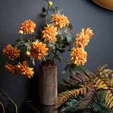 Hill Interiors Orange Dahlia Spray arranged in a textured bronze cylindrical vase against a dark grey wall, with autumnal ferns beside it.