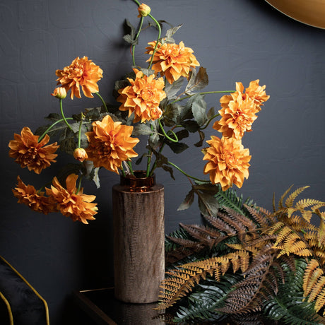 Hill Interiors Orange Dahlia Spray arranged in a textured bronze cylindrical vase against a dark grey wall, with autumnal ferns beside it.