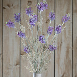 Hill Interiors Purple Meadow Flower stems with clustered ruffled purple blooms and pale green foliage, displayed against a light wooden plank background.