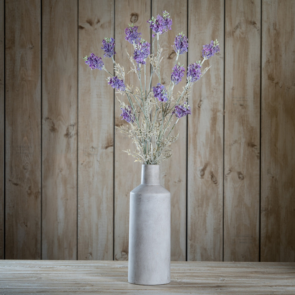 Hill Interiors Purple Meadow Flower faux stems with clustered purple blooms and pale foliage arranged in a tall grey stone bottle vase against wood panelling.