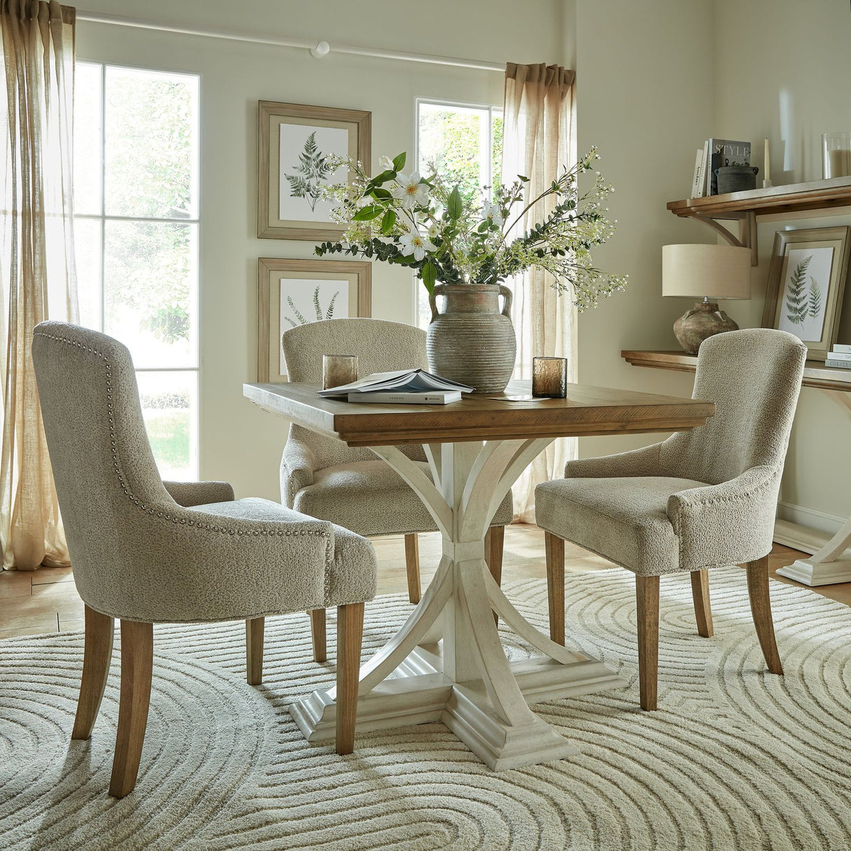 Hill Interiors White Gypsophila Spray arranged with foliage in a stone vase on a round wooden dining table, styled with upholstered chairs in a neutral room.