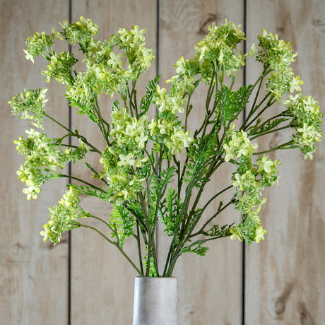 Hill Interiors Green Plumb Blossom Spray faux stem with clusters of small green blossoms and fern-like foliage in a pale vase against wooden panels.