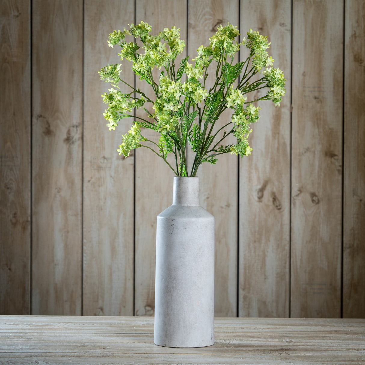 Hill Interiors Green Plumb Blossom Spray in a tall matte grey stone bottle vase on a wooden surface against a rustic timber plank wall.