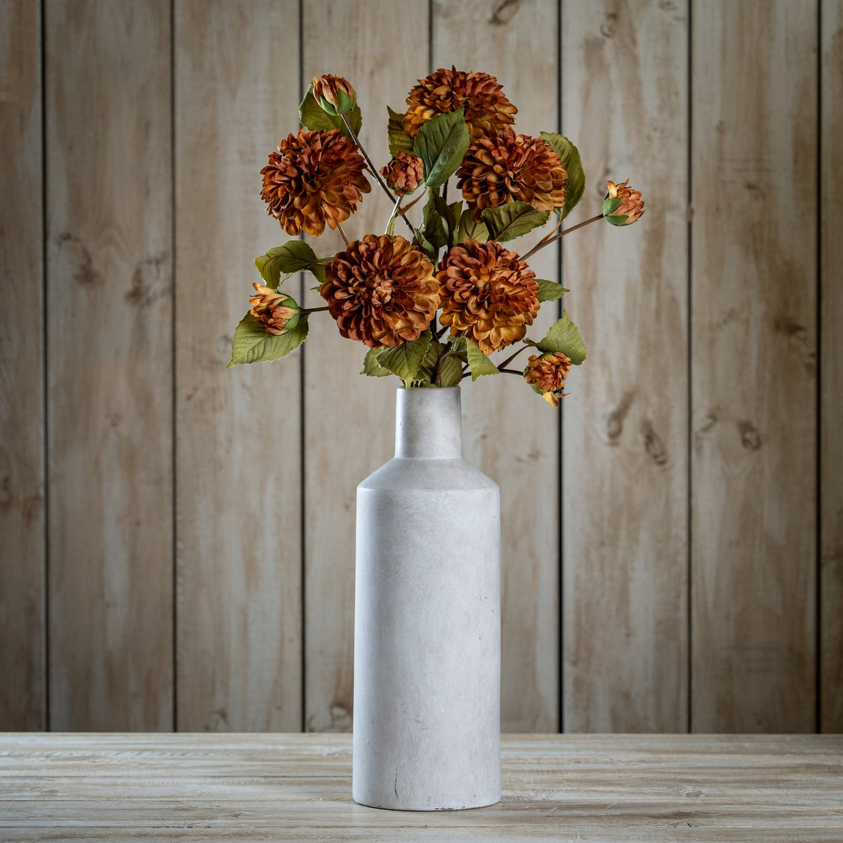Hill Interiors Deep Mustard Dahlia Stem arranged with layered rust blooms and green leaves in a tall matte grey bottle vase against wood panelling.