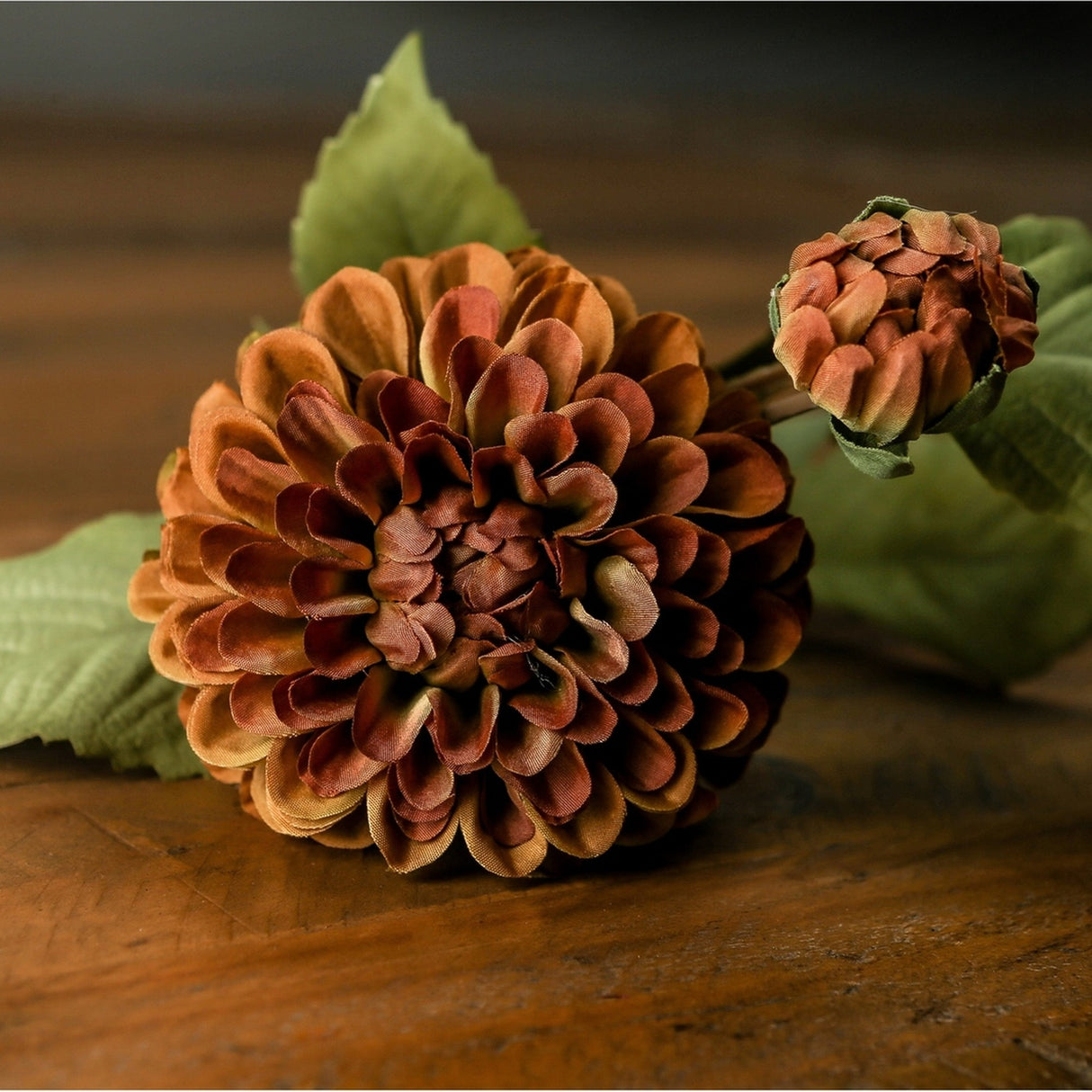 Hill Interiors Deep Mustard Dahlia Stem shown close up with a full rounded bloom, smaller bud and green leaves resting on a dark wooden surface.