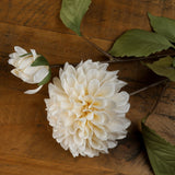 Hill Interiors White Dahlia Stem with a full layered bloom and smaller bud, set against a rustic wood surface with green foliage along the stem.