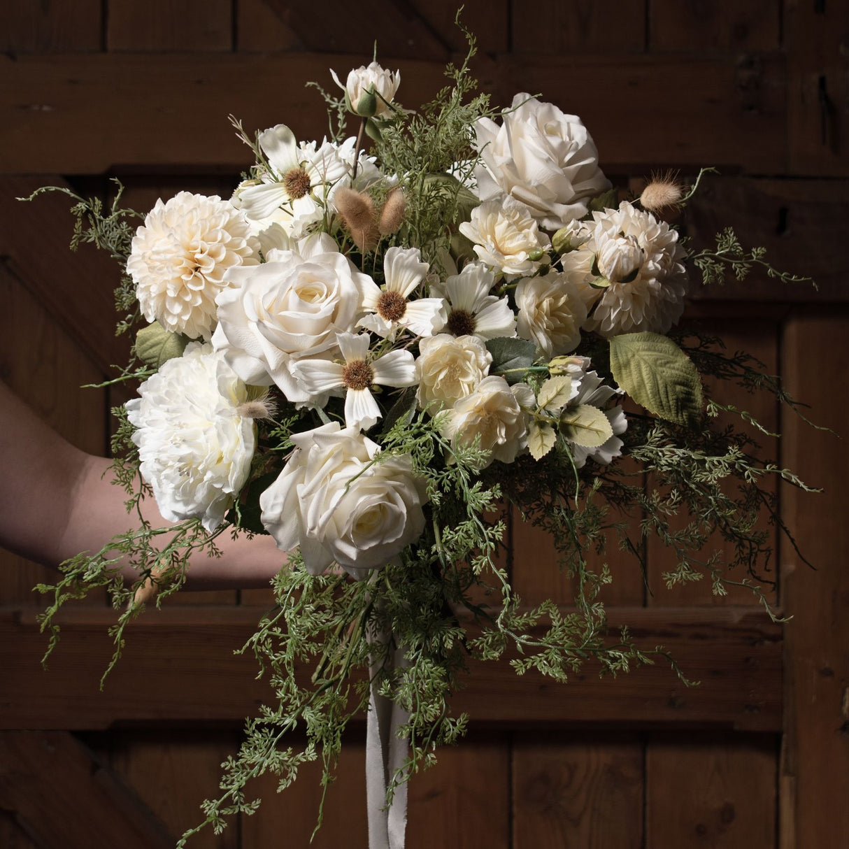 Hand holding a bouquet of white dahlias, roses and cosmos with trailing greenery and bunny tails against a rustic wooden door.