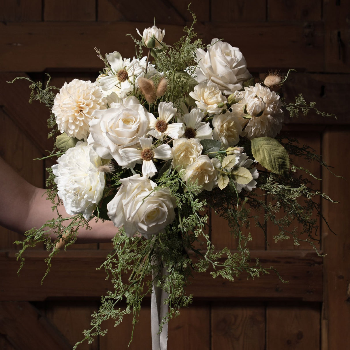 Bridal bouquet featuring Hill Interiors white dahlia stems with faux roses, cosmos and trailing greenery, held against a rustic wooden door.