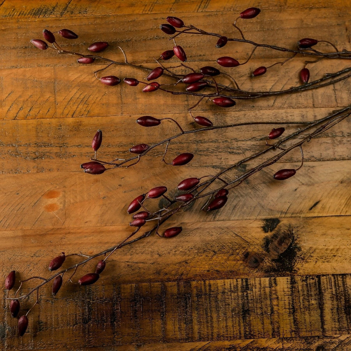 Hill Interiors Burgundy Rosehip Stem faux foliage with clusters of deep red berries on slender branches, laid across a rustic wooden surface.