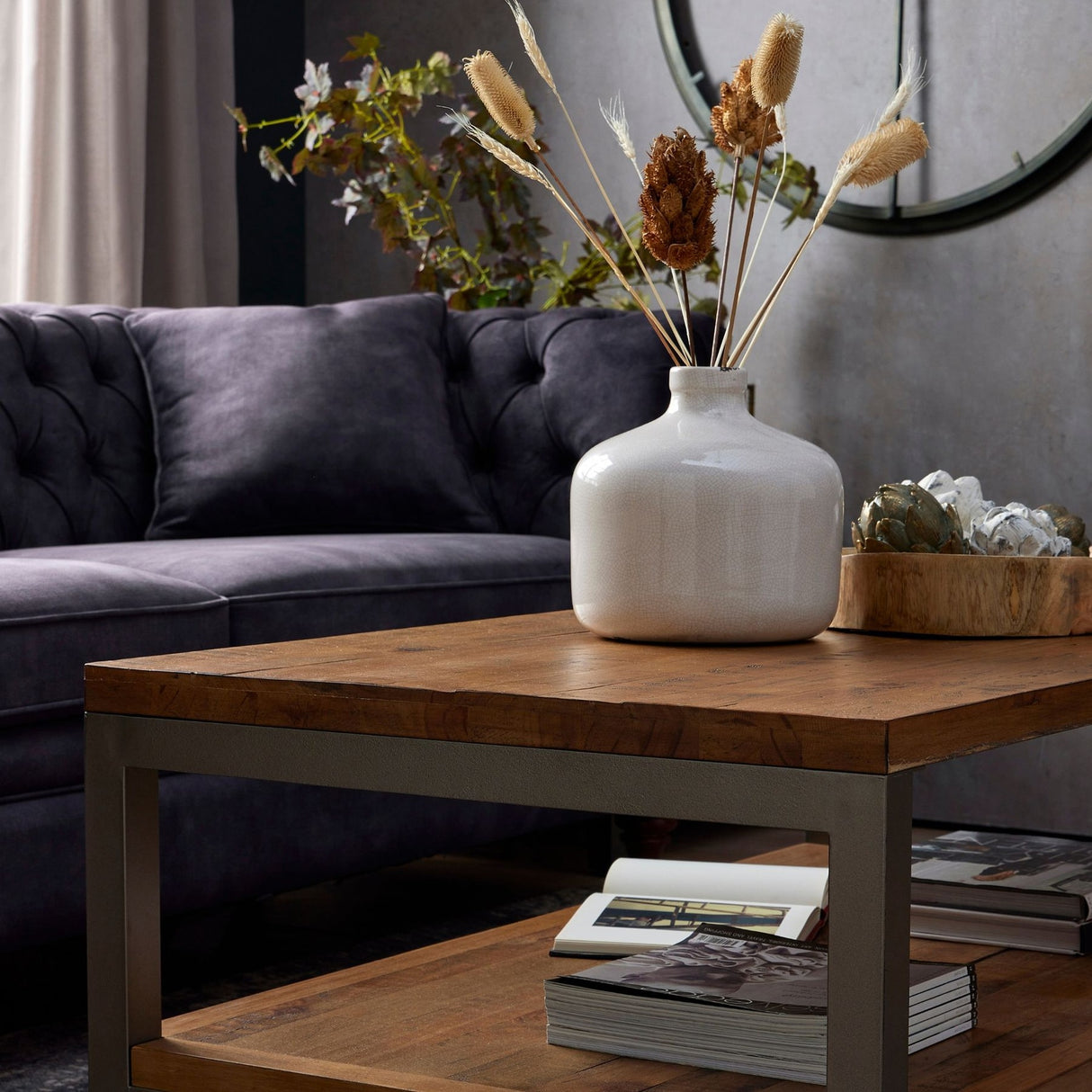 Hill Interiors Bouquet of Dried Tall Thistle in a white ceramic vase on a rustic wood coffee table, styled beside a grey tufted velvet sofa.