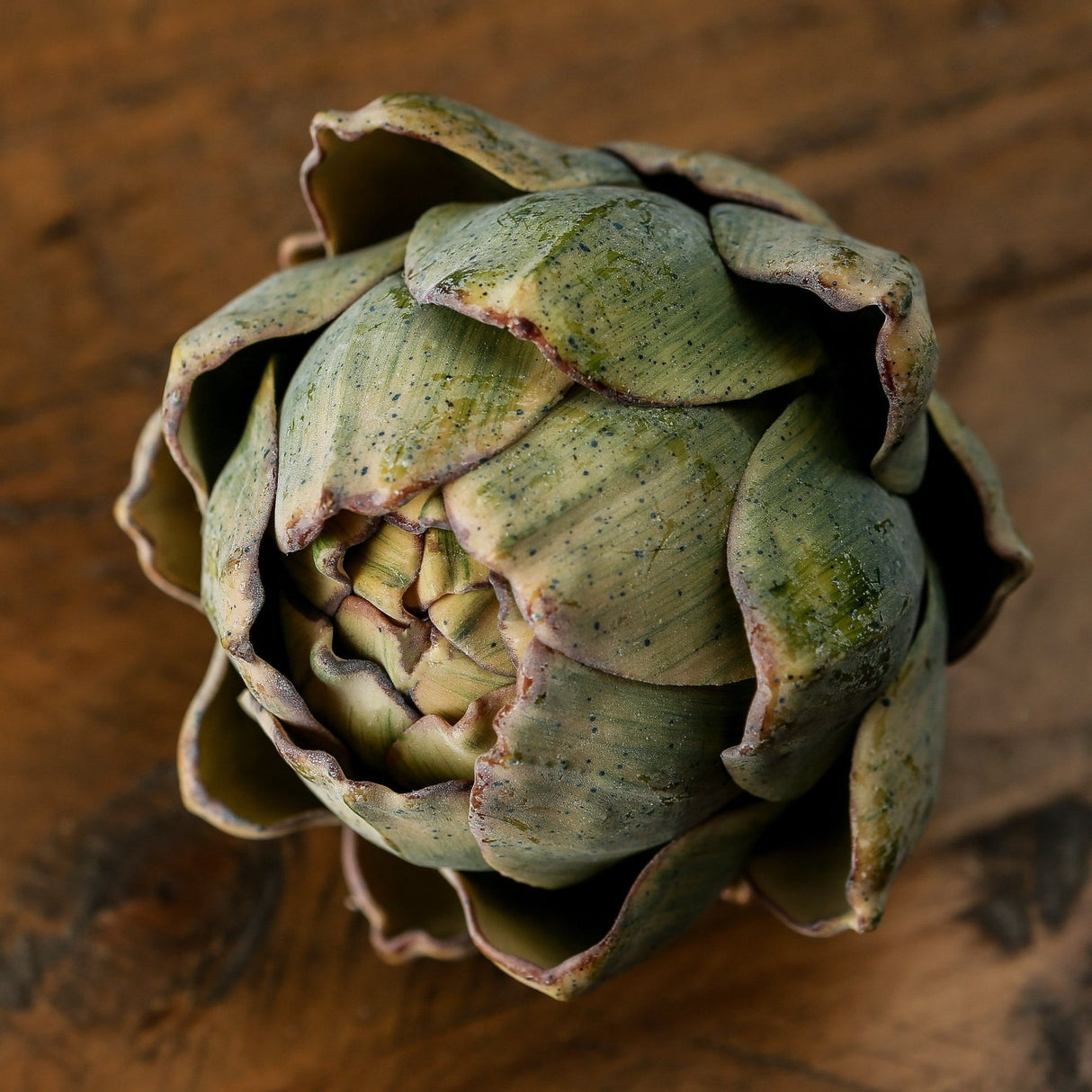 Hill Interiors Real Touch Artificial Artichoke with layered green bracts and tapered points, shown from above on a rustic wooden surface.
