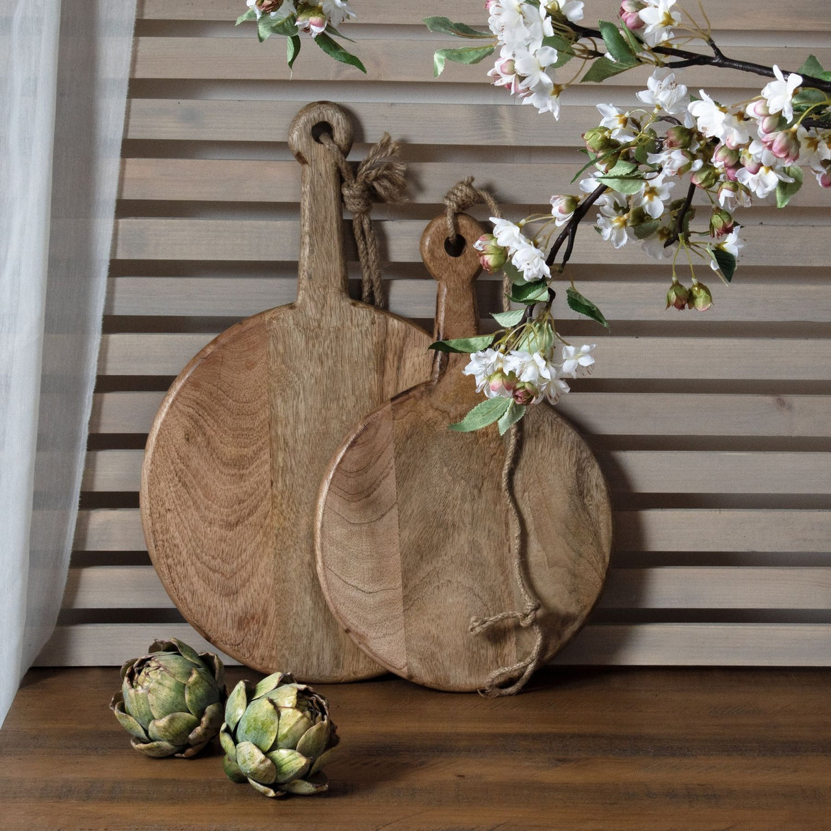 Two Hill Interiors real touch artificial artichokes in graduated green tones displayed beside round wooden boards and blossom stems on a wood surface.