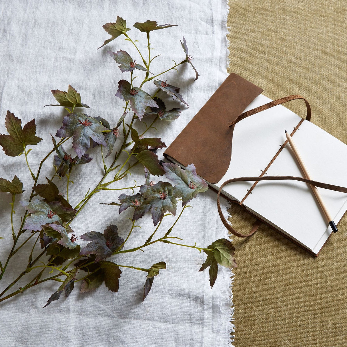 Hill Interiors Variegated Maple Leaf faux foliage stem in green tones, laid on white linen beside a leather-bound journal and pencil on hessian.