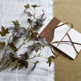 Hill Interiors Variegated Maple Leaf faux foliage stem in green tones, laid on white linen beside a leather-bound journal and pencil on hessian.
