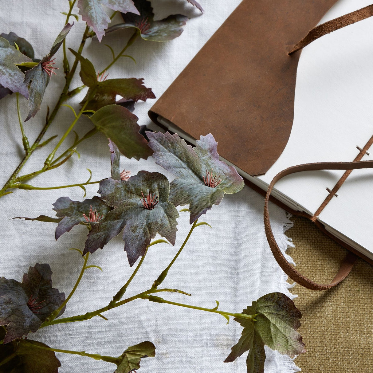 Hill Interiors Variegated Maple Leaf faux foliage stem with green and purple lobed leaves, styled on white linen beside a leather-bound journal.