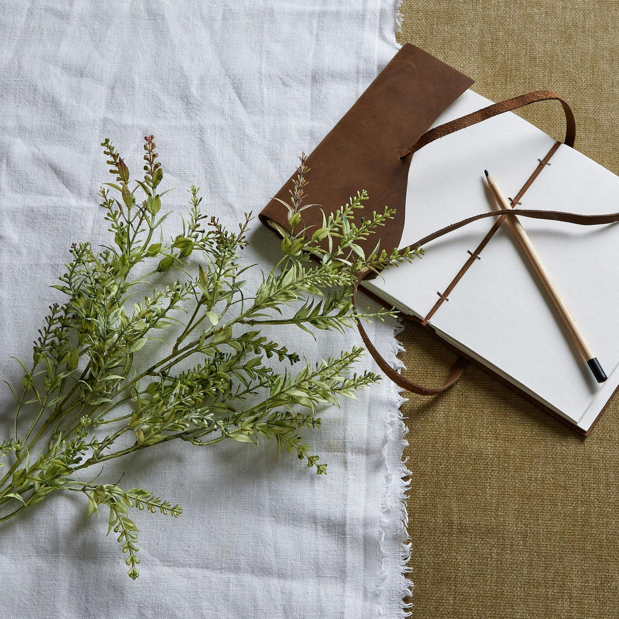 Hill Interiors Broom Stem faux plant with slender green foliage and needle-like leaves, laid on white linen beside a leather-bound journal and pencil.
