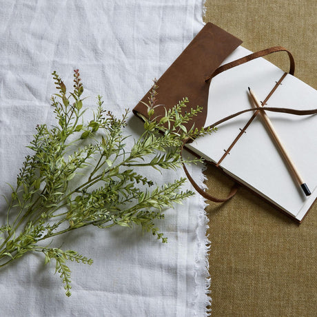 Hill Interiors Broom Stem faux plant with slender green foliage and needle-like leaves, laid on white linen beside a leather-bound journal and pencil.