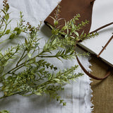 Hill Interiors Broom Stem faux plant with slender green foliage and needle-like leaves, laid on linen beside a brown leather-bound journal.