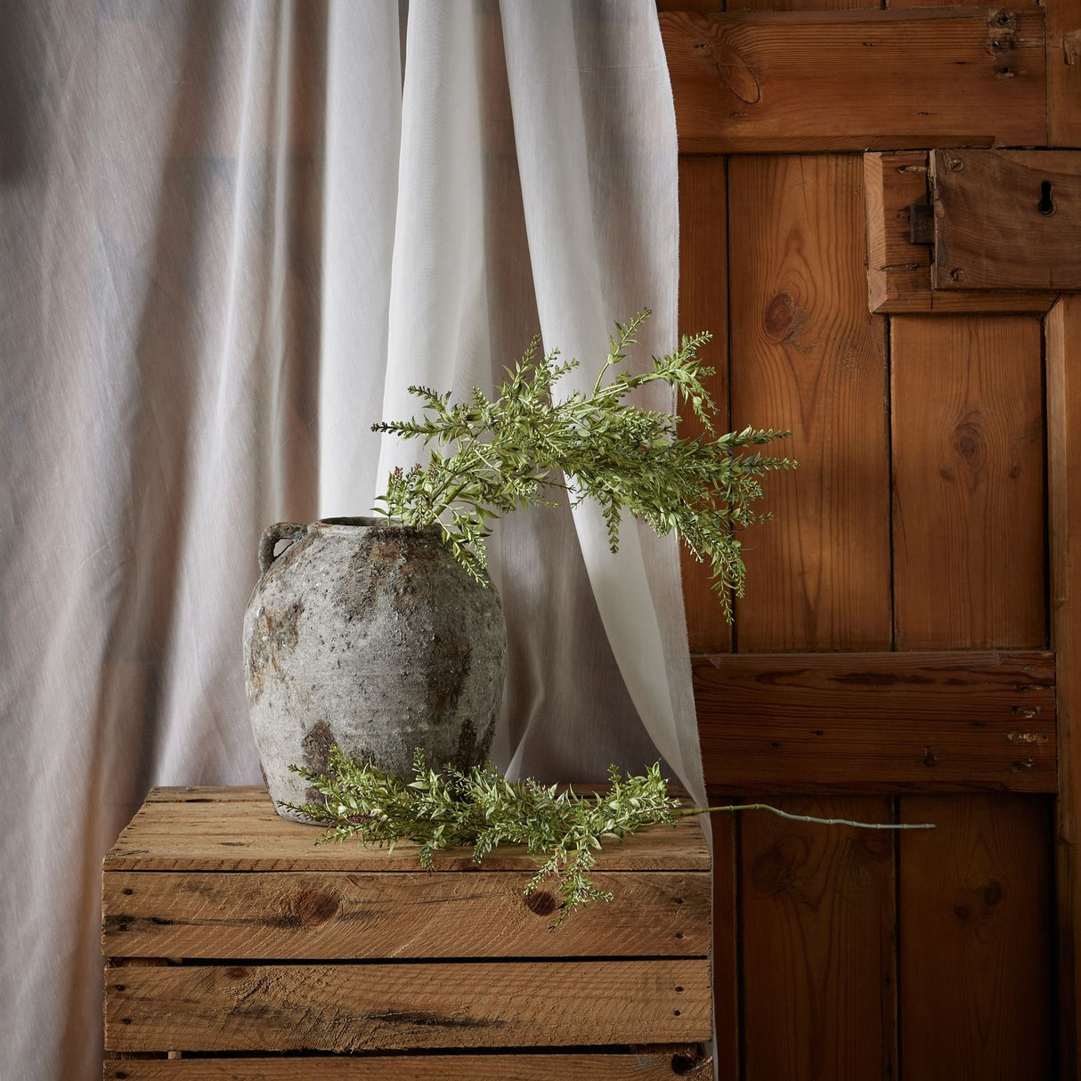 Hill Interiors Broom Stem faux plant with airy green needle-like foliage displayed in a rustic stone jar on a wooden crate beside soft draped linen.