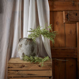 Hill Interiors Broom Stem faux plant with airy green needle-like foliage displayed in a rustic stone jar on a wooden crate beside soft draped linen.