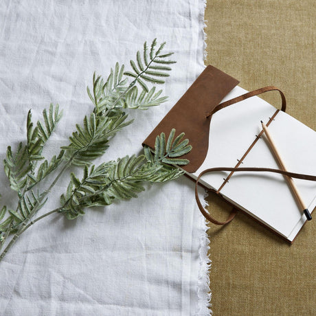 Hill Interiors Silver Wattle Leaf faux foliage stem with feathery fronds laid on white linen beside a leather-bound notebook and pencil.
