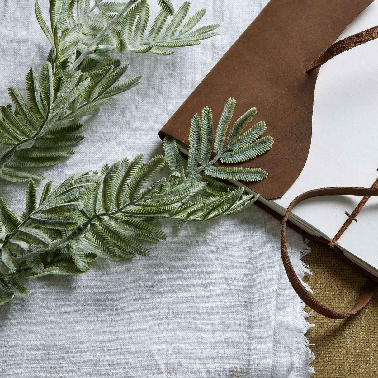 Hill Interiors Silver Wattle Leaf faux foliage stem with feathery silver-green leaflets, draped beside a brown leather-bound journal on white linen.