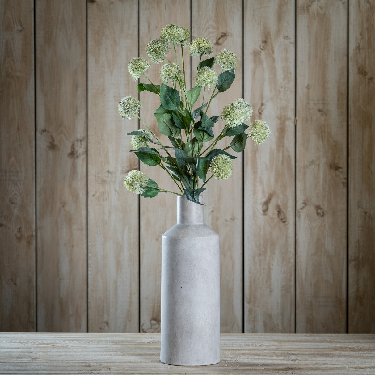 Hill Interiors Thistle Globe Stem with green spiky globe heads and leafy foliage displayed in a tall grey stone bottle vase against a wooden plank backdrop.