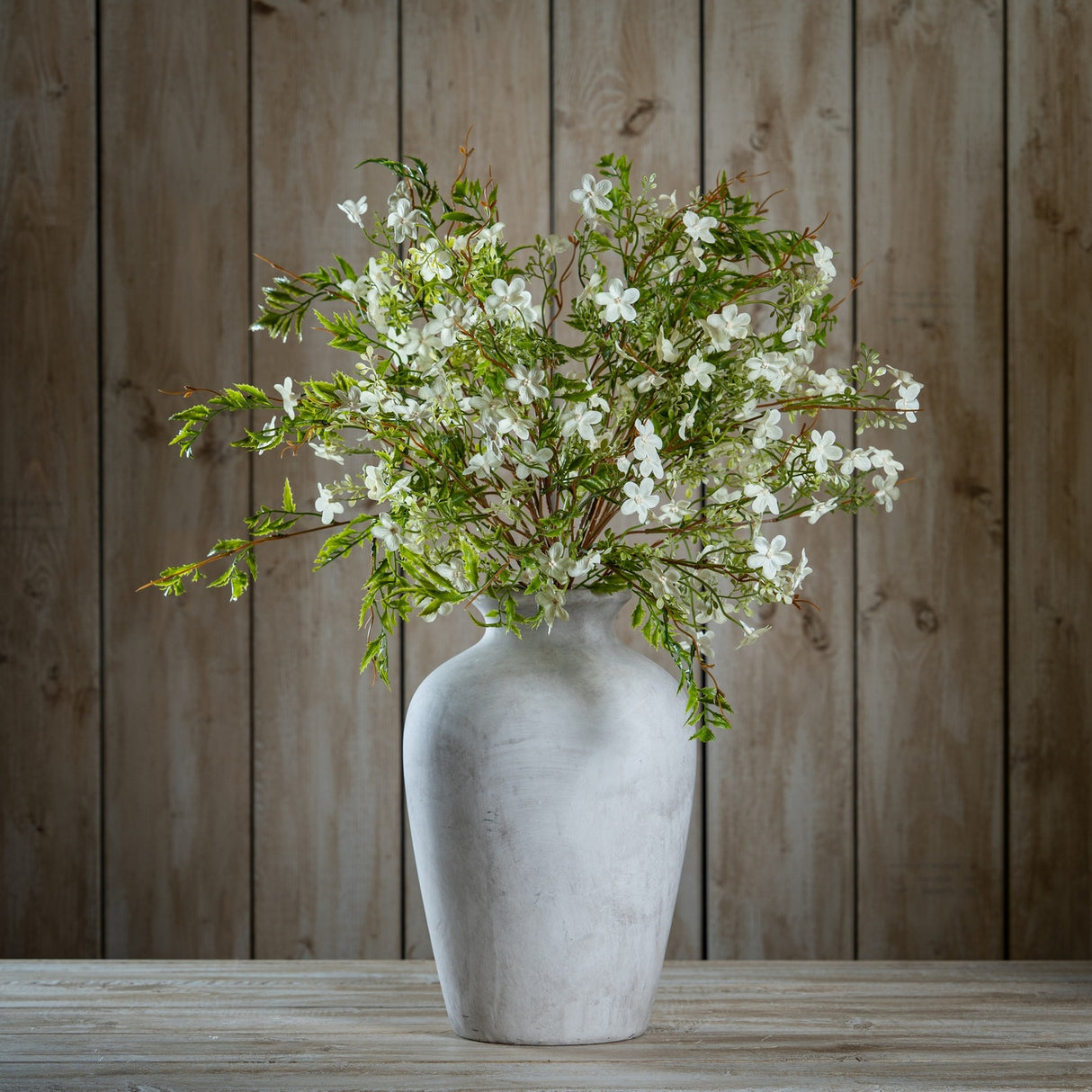 Hill Interiors Italian White Bellflower faux stems arranged in a tall stone vase with green leaves against a wooden plank backdrop.