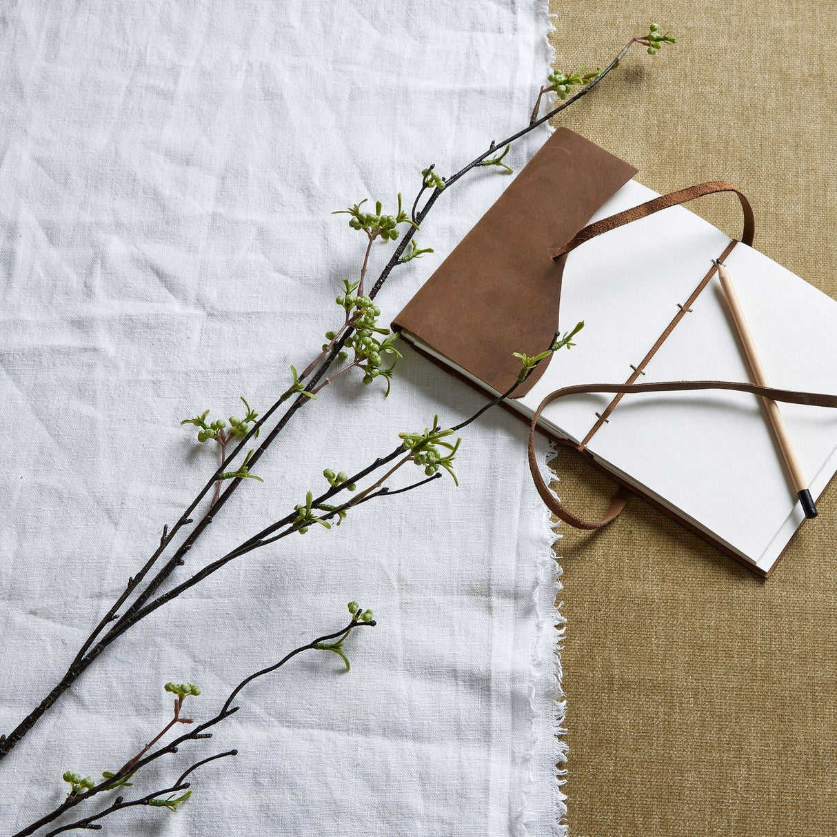 Hill Interiors Green Berry Willow Stem faux foliage with slender leaves and small green berries, arranged on white linen beside a leather journal and pencil.