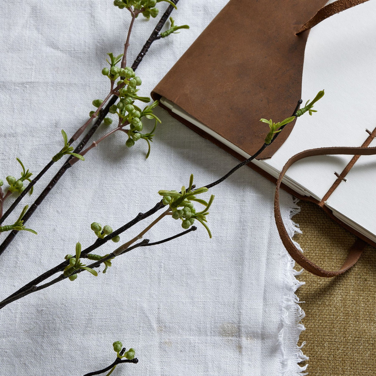 Hill Interiors Green Berry Willow Stem on white linen next to a brown leather journal, showing slender leaves and tiny berries on arching branches.