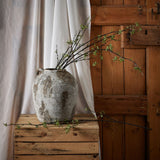 Hill Interiors Green Berry Willow Stems arching from a weathered grey stone jar on a wooden crate, set against linen drapes and a rustic timber door.