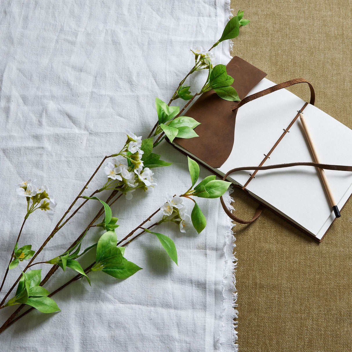 Tall Mock Orange Spray by Hill Interiors laid on white linen beside an open leather-bound journal and pencil, showing white blossoms and green leaves.