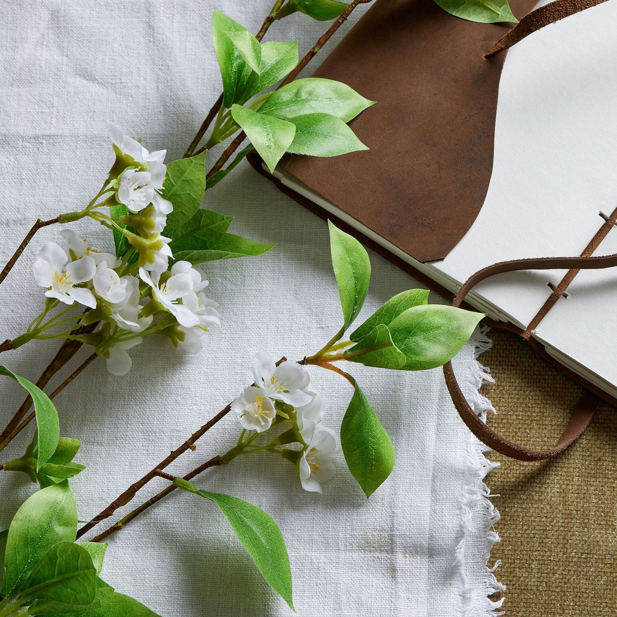 Hill Interiors Tall Mock Orange Spray with white blossoms and green leaves laid beside a brown leather-bound journal on linen fabric.