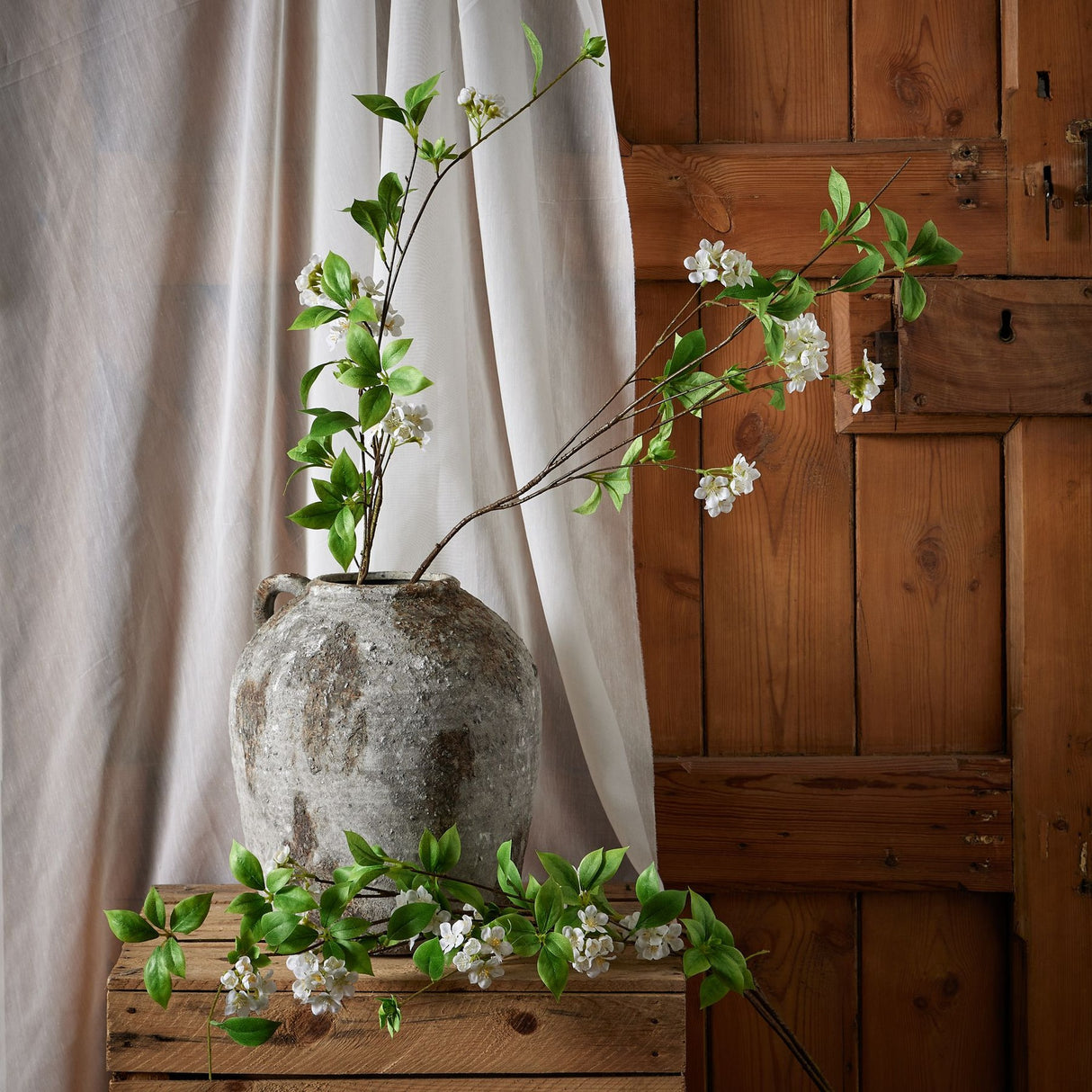 Hill Interiors Tall Mock Orange Spray with white blossoms and green leaves arranged in a rustic stone vase beside a linen curtain and wooden door.